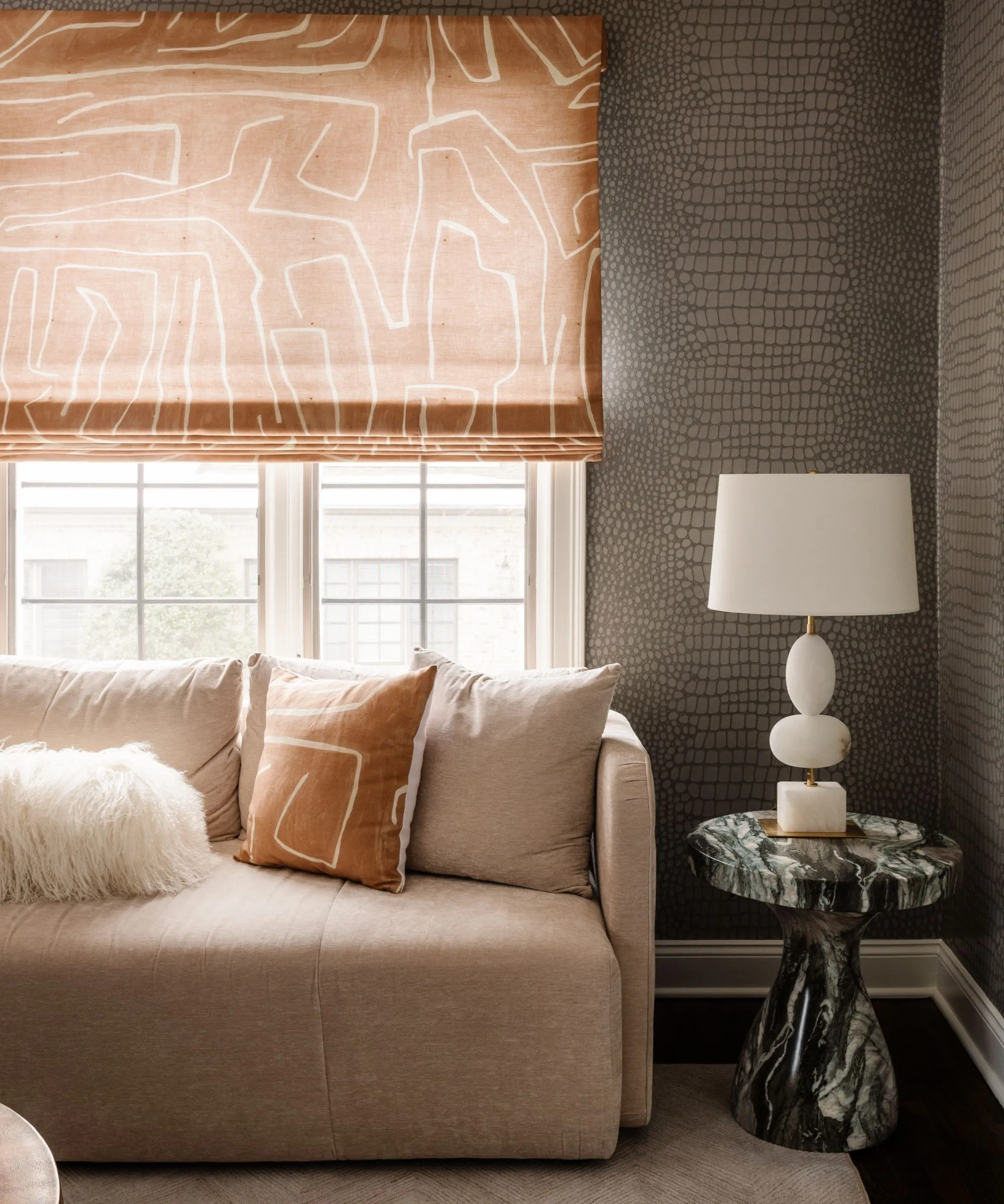 Stue med slangeskindstapet på væggene og et beige sofabord i marmor med hvid lampe og et mønstret orange rullegardin på et vindue over en sofa' loading='lazy' title=