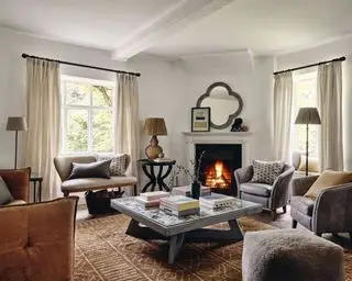 Stor stue med mange sitteplasser, lenestoler og ottomaner, salongbord, peis, to vinduer med gardiner