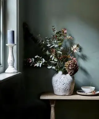 Nærbilde av sesongens blomster og grener i kremkeramikkvase på hylle, blåmalte vegger vinduskarmen med stearinlys i holderen