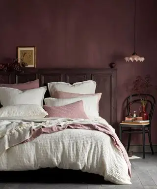 Schlafzimmer mit dunkelrosa gestrichenen Wänden, Blick auf Doppelbett mit weißer Bettwäsche, rosafarbenen Decken und Zierkissen, niedrig hängende Hängelampe über schwarzem Holzstuhl mit Büchern, Blumen und Ornamenten dekoriert, dunkles Holzkopfteil, graubrauner Bodenbelag, gerahmtes Bild und darauf ruhendes Blattwerk am Kopfteil.