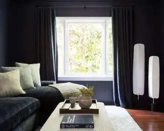 Salón pintado de negro con cortinas negras y sofá, suelo de madera oscura con lámparas de pie blancas estilo farol, alfombra color crema, cojines y reposapiés