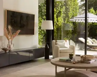 Sala de estar relajada y bohemia, aparador gris bajo con televisor montado en la pared con bordes de madera clara colocados encima, sillón color crema y alfombra natural, mesa de centro escalonada con textura redondeada, tumbonas y sombrilla en el fondo exterior
