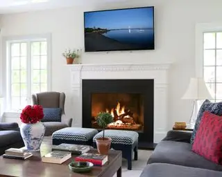 sala de estar blanca y luminosa con una gran chimenea en blanco y negro, sofás y sillón grises, taburetes gemelos con estampado azul marino, mesa de centro de madera oscura, estantería blanca empotrada, televisor montado en la pared encima de la chimenea