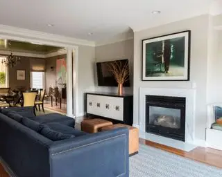 Sala de estar y comedor de planta abierta, pisos de madera con chimenea de mármol, sofá azul con dos taburetes dobles de cuero, asientos tipo alcoba, aparador con televisor montado en la pared arriba y alfombra estampada gris.
