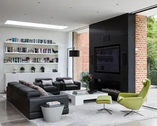 Sala de estar con sofá y sillones de cuero negro, un sillón verde, una mesa de café blanca y un marco de fuego negro con paredes de vidrio a ambos lados
