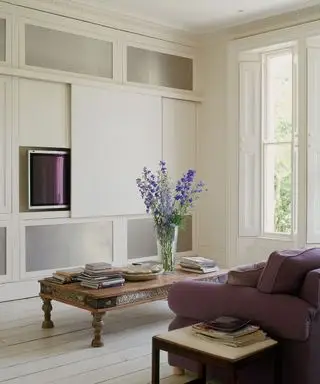 Amplio y moderno salón blanco con suelos encalados, sofá morado, ventanas con contraventanas, pantalla de televisión de plasma oculta tras una pantalla de lona y mesa de centro de madera.