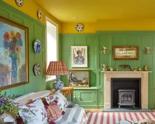 Sala de estar colorida con techo pintado de amarillo, paredes con paneles pintados de verde, mezcla ecléctica de obras de arte y cuadros enmarcados, platos decorativos en la pared, chimenea de piedra tradicional, alfombra a rayas rojas y blancas, mantas a rayas y cojines estampados en el sofá, mesa de cerámica azul y blanca lámpara con pantalla estampada