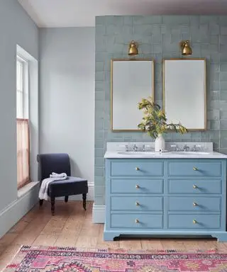 Bagno dipinto di blu con parete piastrellata blu, lavabo blu con due lavandini, due specchi e due lampade da parete, tappeto rosa e sedia nell'angolo della stanza