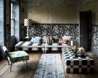 Uferdig gipsvegg og blomstertapet stue, floral pring stol, divansofa i svarte og hvite striper, metallisk rutete ottomanske, skyvevinduer, nakne gulvplanker