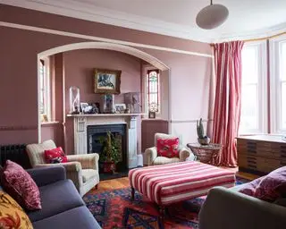 Rosamalt stue med lyst karnappvindu, marineblå sofa, to kremstripete lenestoler, plassert rundt en rosa stripet ottoman, gardiner med matchende rosa stripete mønster, tregulv med flerfarget mønsterteppe, tradisjonell marmorpeis med mantel dekorert med vaser, fotografier og dekorative glassgjenstander