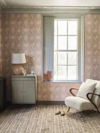 dormitorio con papel tapiz rosa, carpintería gris, sillón tapizado, cómoda pequeña, lámpara y alfombra a rayas