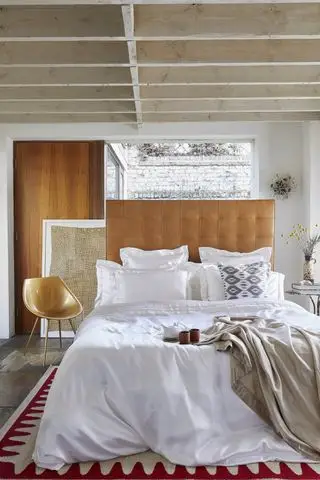 Dormitorio con techo con vigas de madera, suelo de piedra, cabecero de cuero, silla moderna de diseño, ropa de cama de seda blanca, paredes blancas