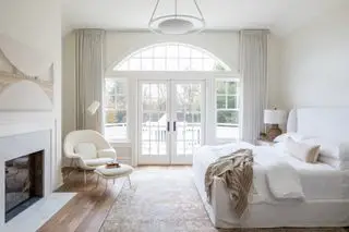dormitorio blanco con silla y reposapiés de boucle, chimenea, arte moderno, alfombra antigua, suelo de madera, vista exterior