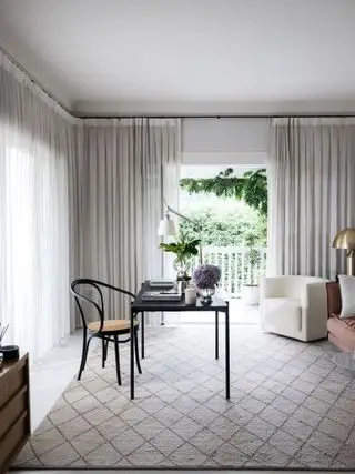 sort skrivebord og stol i stue med mønstret tæppe, cremefarvede gardiner, lænestol, sofa, skrivebordslampe, planter