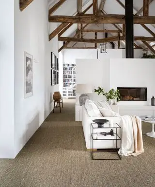 Gran sala de estar blanca de planta abierta, conversión de granero, techo abovedado con vigas de madera oscura a la vista, área para sentarse alrededor de la chimenea, alfombra natural texturizada, vista al área de la biblioteca con escalera blanca