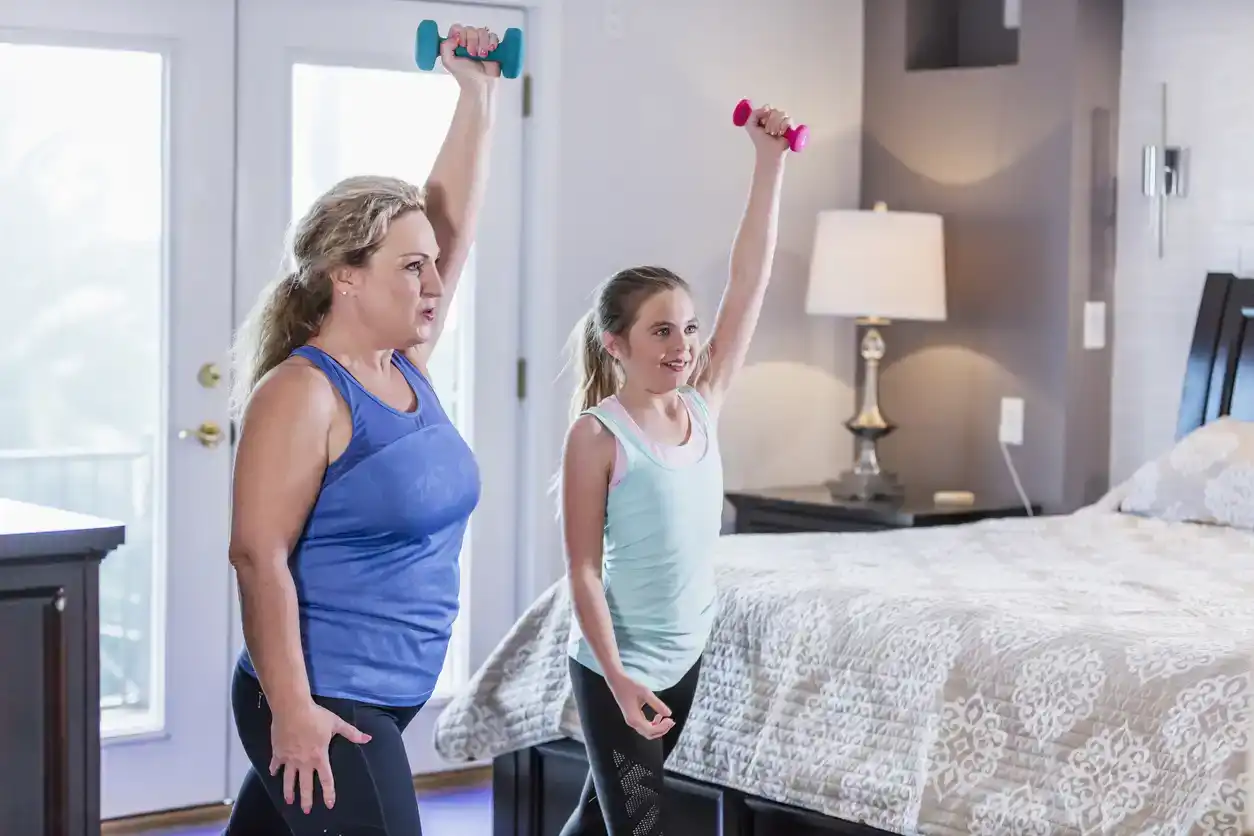 A woman in her 40s and her 11 year old daughter exercising at home together in the bedroom with hand weights, taking an online exercise class