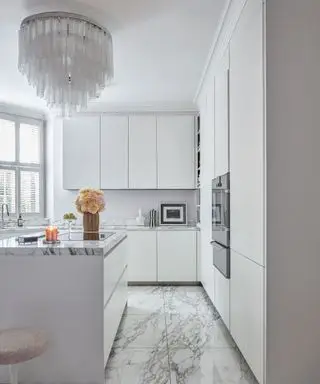 Elegant hvidt køkken med lysekrone i glas, marmorgulv, hvide skabe og køkkenø, lav polstret tulipan skammel