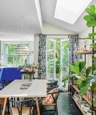 Åbent køkken spisestue, sort sildeben flisegulv, hvidmalede vægge, ovenlysvindue i loftet, lysttræ spisebord med sorte træ spisebordsstole, blomstergardiner, stor reol dekoreret med køkkenudstyr, to hvide hængende pendler over spisebordet