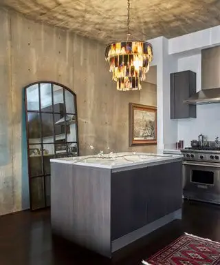 Moderne køkken med væg og loft i betonstil, mørk træø, spejldesign med mørkt træpanel lænet op ad væggen, fjerbeklædt lysekrone over øen