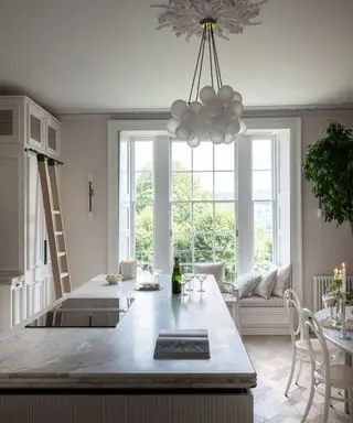 Cuisine blanche élégante avec siège de fenêtre confortable, îlot de cuisine avec dessus en marbre, suspension en verre arrondi, parquet, meubles et armoires blancs
