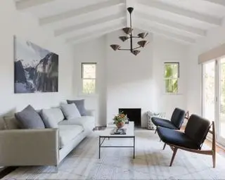 Sala de estar branca com tapete grande, duas poltronas voltadas para sofá cinza, mesa de centro de metal branco e preto, lustre, lareira, obras de arte na parede, teto abobadado