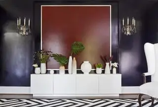 sala de estar violeta con pared roja detrás del aparador, jarrones dispuestos sobre el aparador, alfombra monocromática y apliques estilo candelabro adornados de época. Sillón blanco