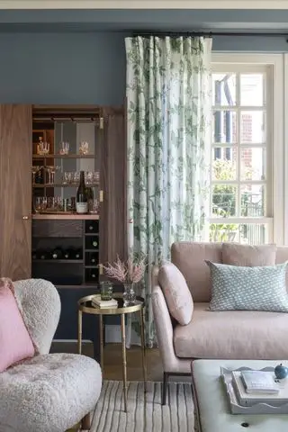 sala de estar azul con cortinas florales verdes, sofá rosa, mesa auxiliar dorada, mueble bar, reposapiés y alfombra texturizada