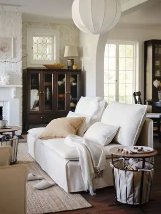 Sala de estar blanca y neutra con sofá blanco de estilo moderno, mesas auxiliares de madera y metal, alfombra neutra, cojines color topo y expositor estilo caoba.