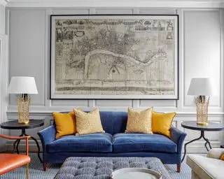 Sala de estar com paredes em painéis cinza claro. sofá azul com almofadas amarelas e mesinhas laterais redondas e estampa de mapa.