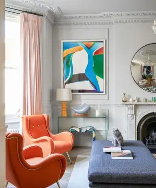 Sala de estar pintada de cinza com painéis, pintura colorida emoldurada, cortinas rosa e poltronas gêmeas laranja, mesa de console de vidro e lareira de mármore, grande pufe azul,