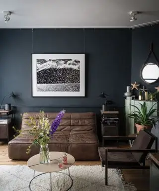 Sala de estar pintada em azul escuro com sofá e poltrona de couro marrom, tapete creme sobre piso de madeira clara, mesa de centro arredondada, lareira de azulejos verdes claros