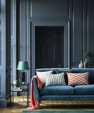 Sala pintada de azul com painéis, sofá azul com almofadas coloridas e manta rosa, mesa lateral metálica com abajur azul, piso de madeira clara