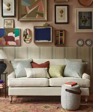 Sala com painéis de tecido listrado, paredes pintadas em rosa claro com parede de galeria ou quadros e objetos decorativos emoldurados, tapete colorido e repousa-pés arredondado, sofá creme de dois lugares com almofadas espalhadas coloridas