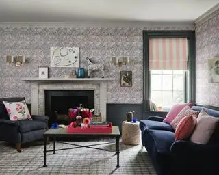 Sala com papel de parede dado atelier, painéis pintados de azul e detalhes em madeira, sofá e poltrona azuis, almofadas rosa e vermelhas, mesa de centro em metal preto, lareira em mármore, persiana listrada rosa e branca, luminárias de parede com dois abajures e quadros de parede