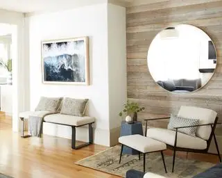 Sala com painéis de madeira aparente e grande espelho redondo montado na parede, paredes pintadas de branco com rodapé pintado de bege, poltrona e banco creme e preto com almofadas de assento, piso e tapete de madeira clara, mesa lateral azul e arte de parede