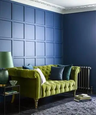 Sala pintada de azul com painéis quadrados, sofá de dois lugares em veludo verde, piso preto e tapete cinza, mesa lateral preta metálica com abajur verde