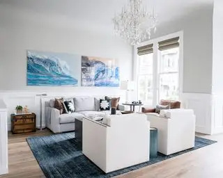 Kyststue med to kunstværker på væggen, lysegrå malet væg, stor glaslysekrone, blåt tæppe over trægulv, to creme lænestole mod en grå sofa med mørkegrå sofabord