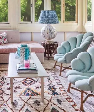 Stue med creme-tæpper og creme- og flerfarvet tæppe, to lyseblå, polstrede lænestole med tunge lænestole vendt mod et lyst træ, rektangulært sofabord, hjørne af en pink mønstret daybed, dekorativ, mønstret lampeskærm på mørkt træ sidebord