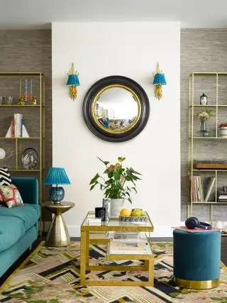 Sala de estar con paredes blancas, espejo circular llamativo con apliques a ambos lados. Mesa de centro dorada, alfombra llamativa y sofá y reposapiés azules