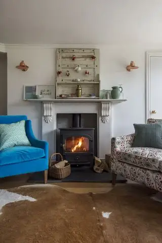 Salón con sillón de terciopelo azul junto a la chimenea con paneles de madera desgastados y alfombra de piel de animal debajo