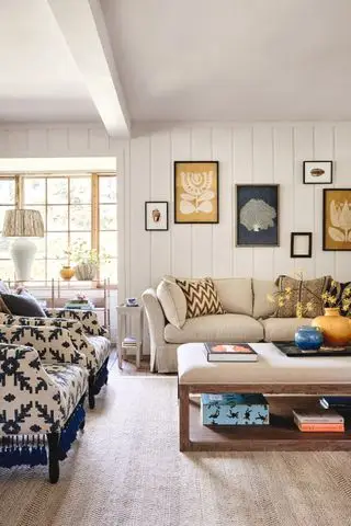 stue med hvite shiplap vegger, beige sofa og polstret salongbord, teppe, marine og beige print lenestoler, kunstverk
