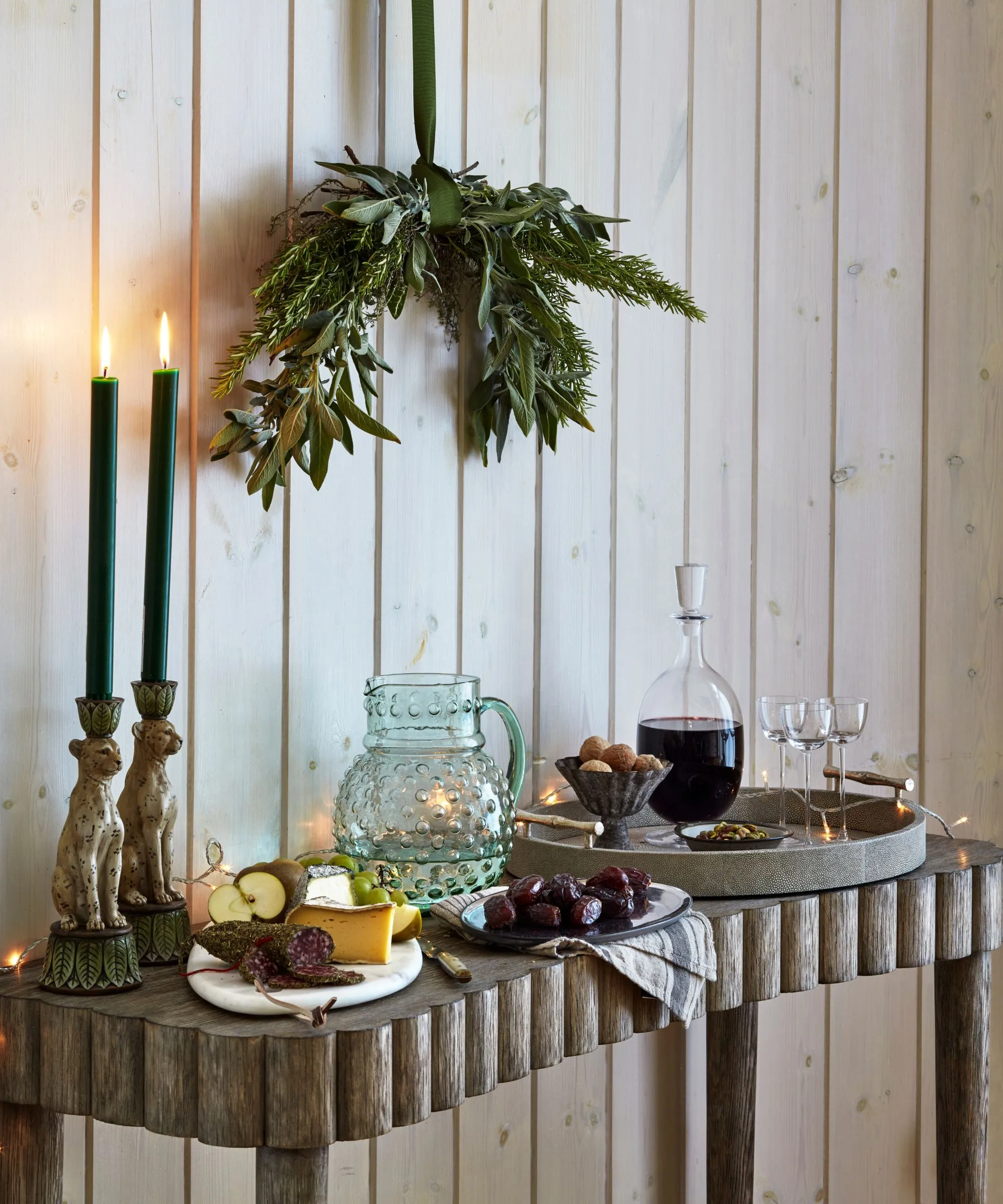 Tavolo da console in legno set con un decanter di vino e bicchieri una brocca di acqua e candele rastremate con un mucchio di fogliame appeso sopra' loading='lazy' title=