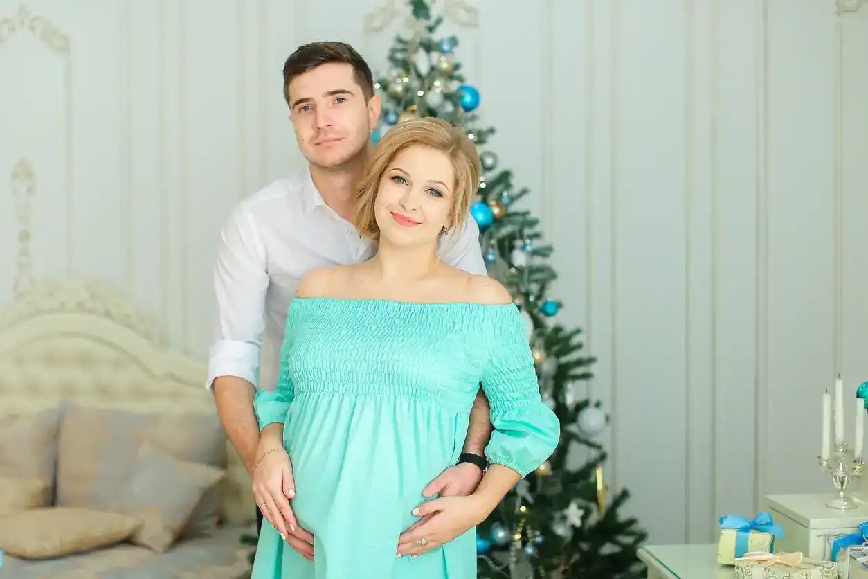 Pregnant caucasian woman wearing blue dress standing with husband near Christmas tree