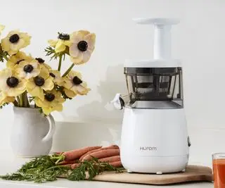 Exprimidor lento Hurom sobre una tabla de cortar con zanahorias y un jarrón de flores en el fondo