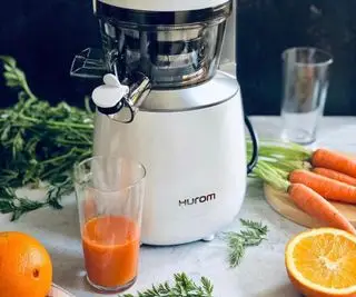 Exprimidor lento Hurom en blanco sobre una encimera con zanahorias y naranjas alrededor