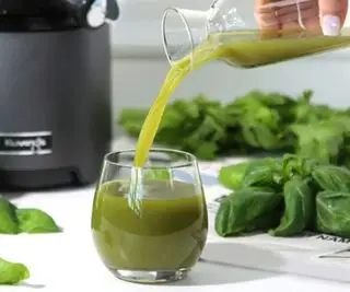 Jugo verde vertido en un vaso con hojas y el exprimidor Kuvings al fondo