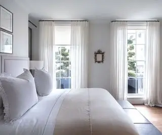 Letto matrimoniale in una camera da letto grigio chiaro con pavimento in legno, biancheria da letto color crema e bianco e tende bianche che vestono finestre dal pavimento al soffitto. Una residenza londinese ristrutturata di quattro piani, casa di Louise Bradley, designer d'interni.