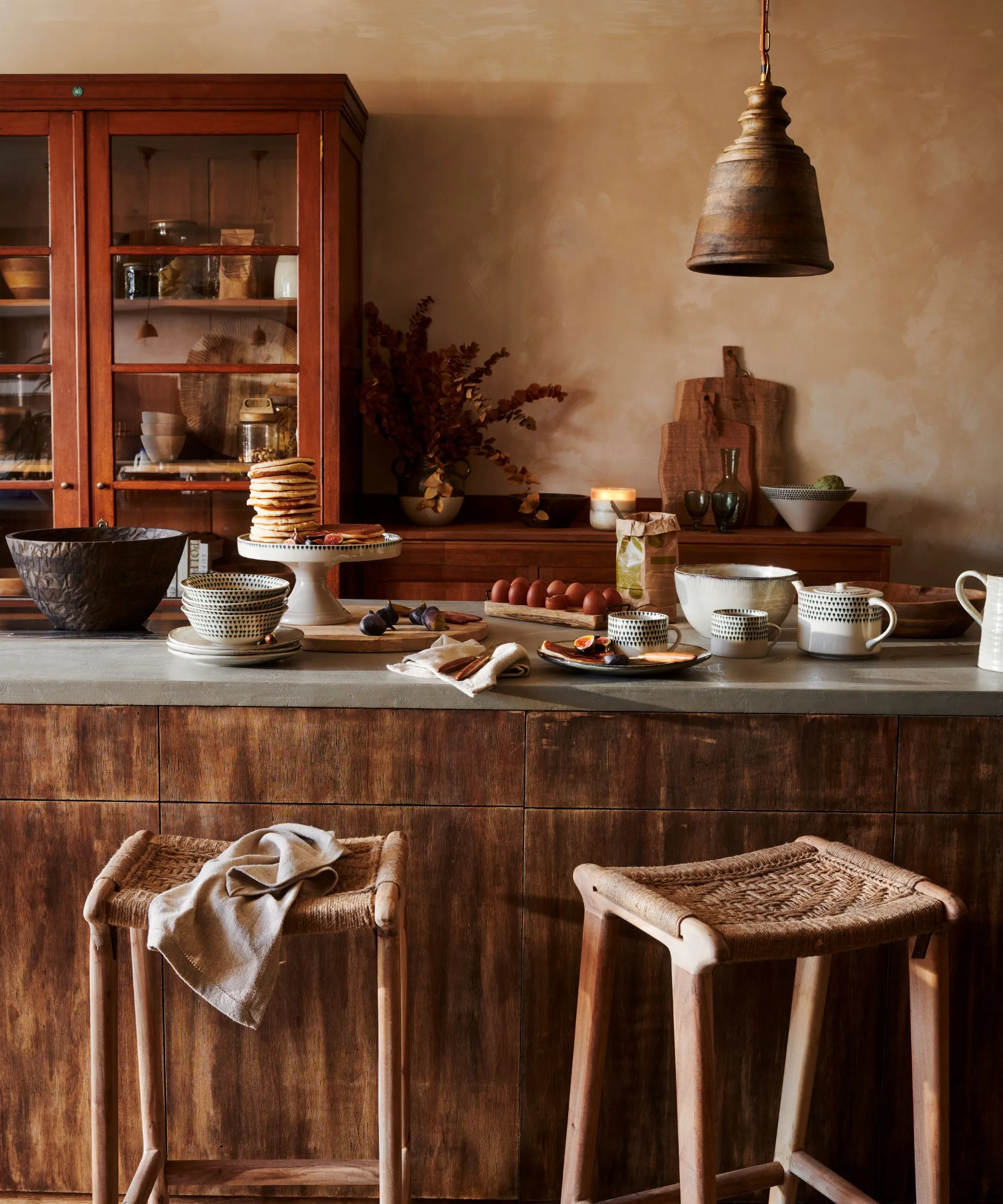 Una cucina dai toni della terra con isola in legno e dispensa dipinta di rosso. I pancake sono impilati su un'alzatina sull'isola. Si avvicinarono due sgabelli di rattan.' loading='lazy' title=