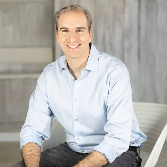 et bilde av den profesjonelle arrangøren Ben Soreff satt på en hvit stol mot en trebakgrunn iført en lyseblå langermet skjorte med krage og svarte jeans' loading='lazy' title=