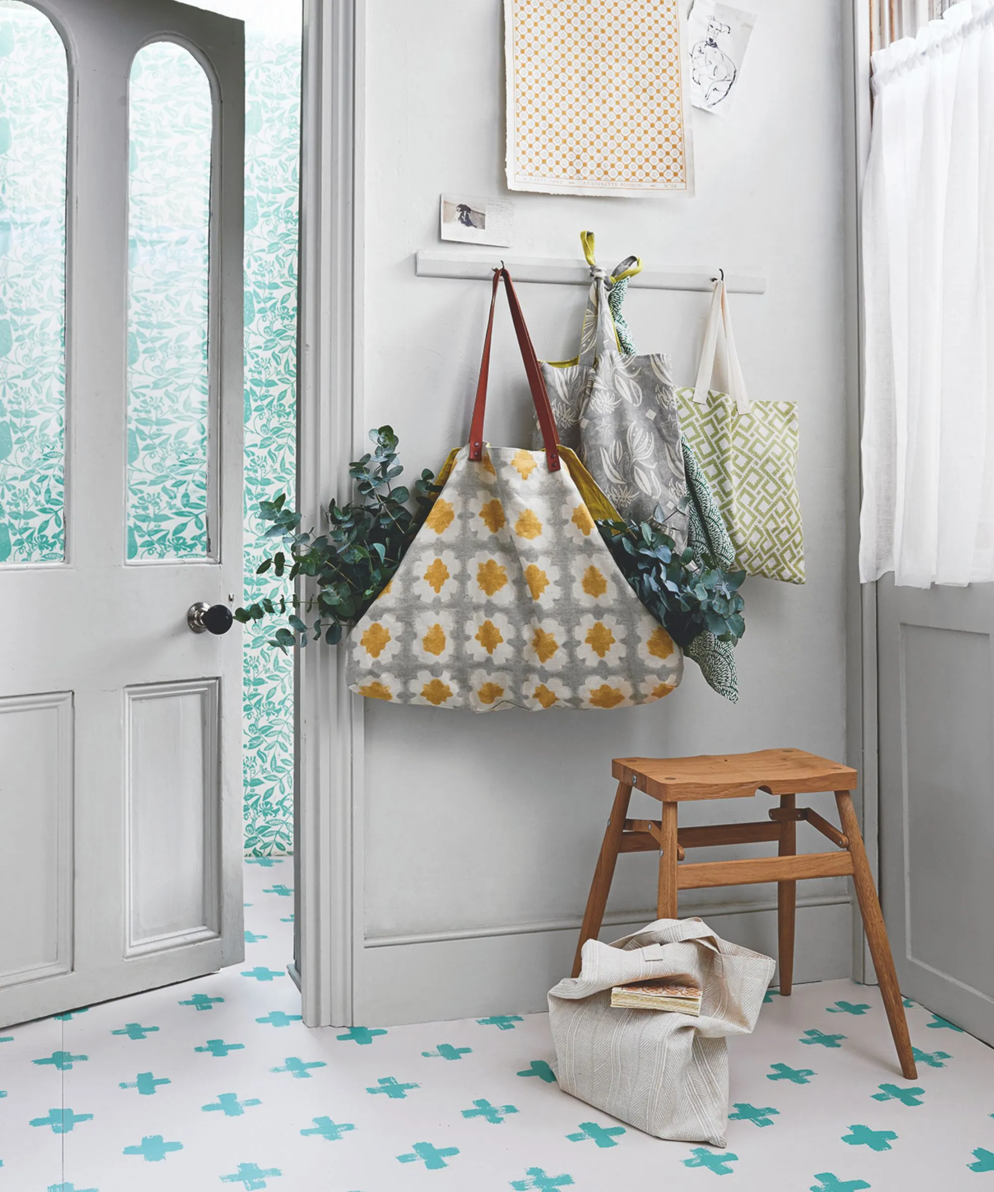 En hvit entré med blå og hvite fliser på gulv, en hvit glassdør og veggkroker med poser fylt med planter. Under dem er en trekrakk og til høyre er et vindu med en gjennomsiktig gardin.' loading='lazy' title=